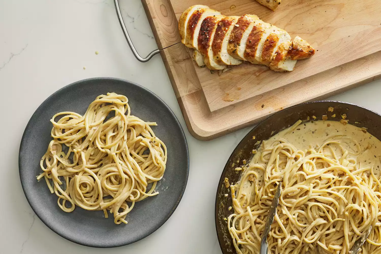 A small plate with creamy pasta, with a cutting board of sliced chicken breast, next to a skillet of more pasta tossed in cream sauce