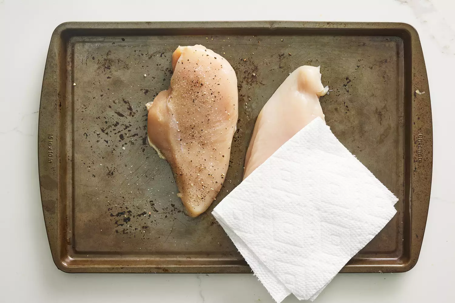 A chicken breast topped with salt and pepper, and a chicken breast with a paper towel on top